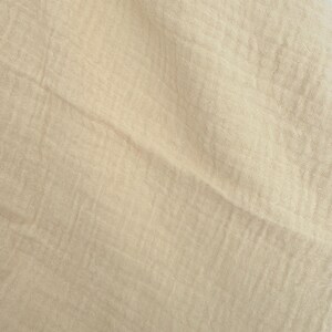 Puede incluir: Primer plano de una tela beige con un aspecto texturizado y arrugado. La tela tiene un color suave y apagado y un tejido ligeramente irregular, lo que sugiere un textil de fibra natural. Los pliegues y las sombras crean una sensaci&oacute;n de profundidad y dimensi&oacute;n.