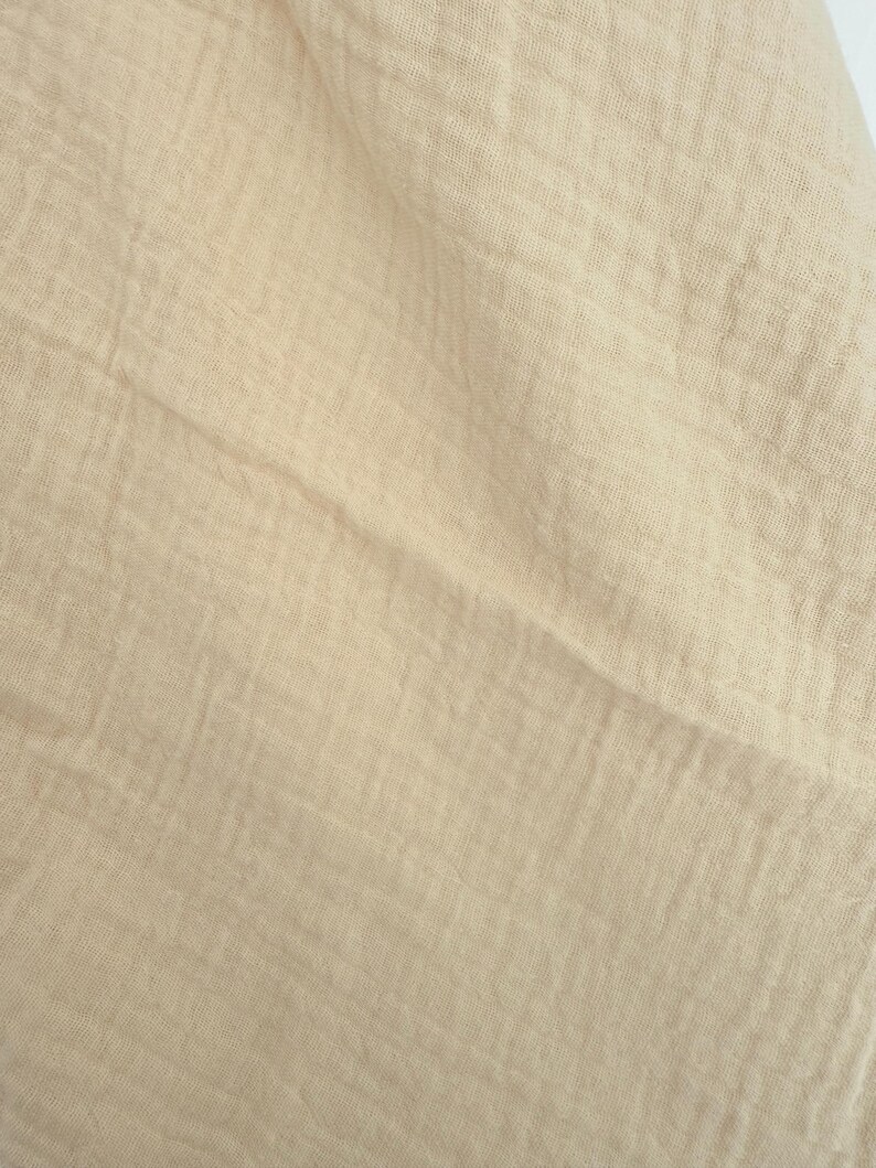 Puede incluir: Primer plano de una tela beige con un aspecto texturizado y arrugado. La tela tiene un color suave y apagado y un tejido ligeramente irregular, lo que sugiere un textil de fibra natural. Los pliegues y las sombras crean una sensaci&oacute;n de profundidad y dimensi&oacute;n.