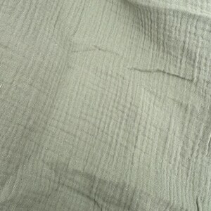 Puede incluir: Primer plano de una tela verde salvia con una apariencia texturizada y arrugada. La tela tiene una ca&iacute;da suave y fluida, con l&iacute;neas verticales y horizontales visibles. El material parece ligero y transpirable, adecuado para ropa o decoraci&oacute;n del hogar.