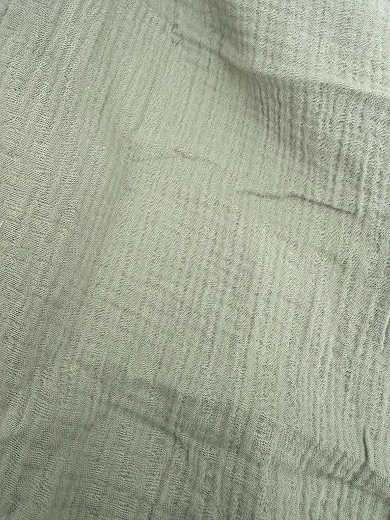 Puede incluir: Primer plano de una tela verde salvia con una apariencia texturizada y arrugada. La tela tiene una ca&iacute;da suave y fluida, con l&iacute;neas verticales y horizontales visibles. El material parece ligero y transpirable, adecuado para ropa o decoraci&oacute;n del hogar.