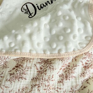 Peut inclure: Une couverture pour bébé blanche avec un motif floral et le nom "Diane" en écriture noire. La couche supérieure présente un motif texturé à pois, et les bords sont bordés d'un tissu rose clair.