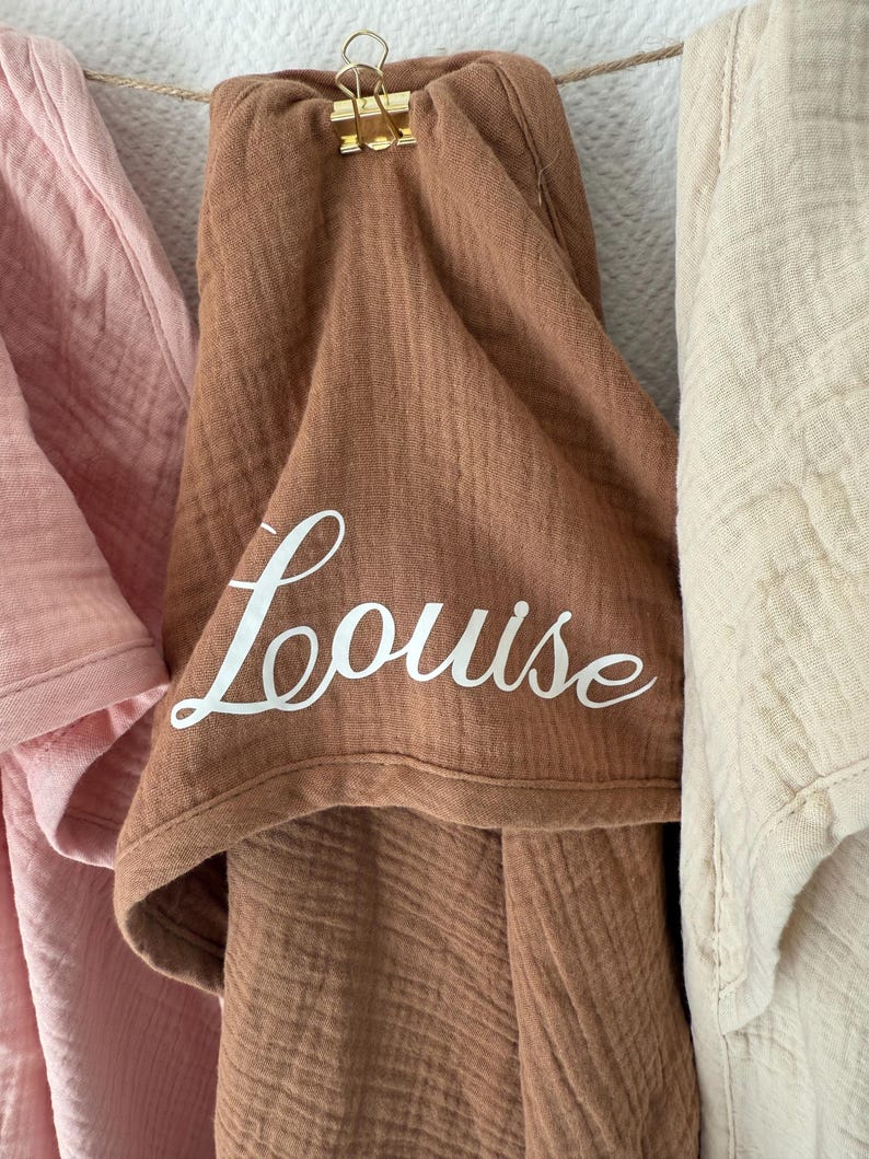 Puede incluir: Una muselina marr&oacute;n con el nombre "Louise" en escritura blanca est&aacute; sujeta a un tendedero con un clip dorado. A cada lado, una muselina rosa y otra crema.