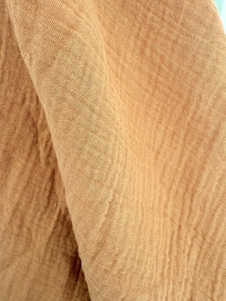 Puede incluir: Primer plano de una tela plegada y texturizada en un tono c&aacute;lido y terroso. La tela tiene un aspecto arrugado, con hilos tejidos visibles. El color es un marr&oacute;n claro apagado, que recuerda al lino o al algod&oacute;n natural.