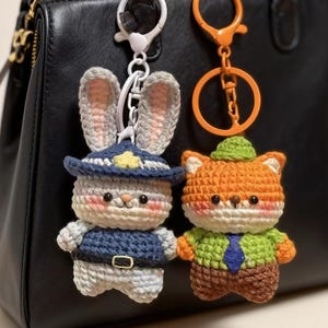 Pode incluir: Chaveiros de crochê de um coelho e uma raposa. O coelho usa um chapéu e uniforme de policial azuis. A raposa está vestida com uma jaqueta verde e calças marrons. Ambos têm chaveiros. Os chaveiros estão pendurados em uma bolsa preta.