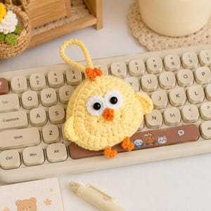May include: A yellow crocheted chick pouch with a loop handle, featuring large black and white eyes, an orange beak, and small orange feet. The circular yarn accessory is positioned on a beige computer keyboard with rounded keys and a spacebar.