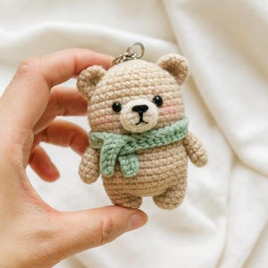 以下が含まれることがあります： ベージュ色の糸で編まれたクマのキーホルダー。緑色のスカーフを巻いています。クマは黒い目、黒い鼻、ピンクの頬をしています。キーホルダーは銀色の金属リングで吊り下げられています。