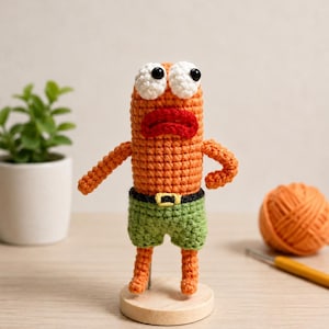 May include: A crocheted orange character figure with large white eyes, black pupils, and a red mouth. The figure wears green shorts with a black belt and a yellow buckle. It stands upright on a small, round, light-colored wooden base.