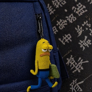 Puede incluir: Llavero de ganchillo amarillo y azul que representa un personaje de dibujos animados con una expresión triste. El llavero está sujeto a una mochila azul marino. El personaje sostiene un objeto verde.