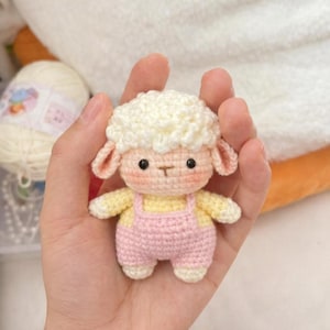 Op de afbeelding: Een kleine, handgemaakte gehaakte schaapjespop met een pluizige witte kop, een roze tuinbroek en een geel shirt. Het schaap heeft kleine, roze oren en een schattig, lachend gezicht. De pop wordt in een hand gehouden.