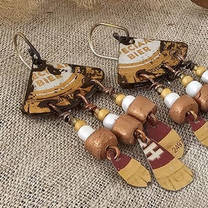 Peut inclure: Boucles d'oreilles artisanales composées de capsules de bière recyclées avec l'inscription "ECIAA BIER". Elles présentent un design original avec des perles cuivrées et des éléments pendants, pour un look rustique et artistique. Fond en lin neutre.