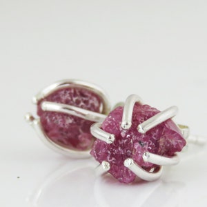 Op de afbeelding: Een paar oorknopjes met ruwe, dieproze edelstenen gezet in een zilverkleurige metalen kooi. De oorbellen worden gepresenteerd op een wit oppervlak, waardoor het licht wordt gereflecteerd.