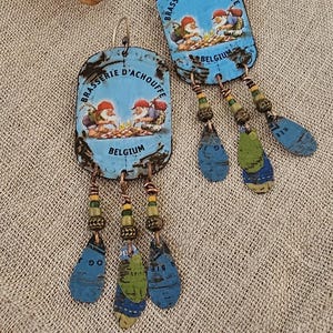 Pode incluir: Brincos artesanais com um design retangular azul e o texto "Brasserie d'Achouffe Belgium". Os brincos têm uma franja de contas pendurada em tons de azul, verde e dourado. Colocados sobre uma superfície de linho bege.