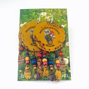 Könnte beinhalten: Handgefertigte Ohrringe aus recycelten Bierdeckeln. Die gelben Deckel zeigen den Text "BRASSERIE D'ACHOUFFE" und "BELGIUM". Die Ohrringe haben bunte Perlen und Anhänger in verschiedenen Formen und Größen.