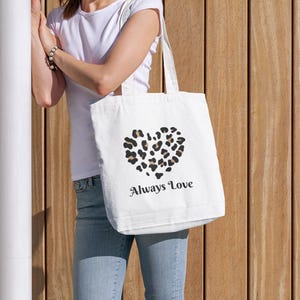 Könnte beinhalten: Eine weiße Tragetasche mit einem herzförmigen Leopardenmuster und den Worten "Always Love" darunter. Die Tasche wird von einer Person getragen. Der Hintergrund ist eine Holzwand.