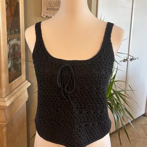 May include: Black crochet tank top with a scoop neckline and tie detail. The top is displayed on a mannequin. The open weave design is visible. The tank top is a summer fashion item.