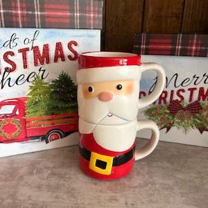 May include: A ceramic mug shaped like Santa Claus, featuring a red hat rim, a white beard, a black belt with a yellow buckle, and a handle on the side. The mug is positioned in front of two decorative boxes with red plaid patterns and Christmas-themed text.