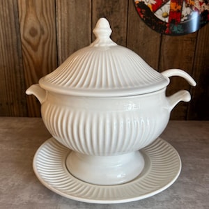 May include: A white ceramic soup tureen with a lid, handles, and a matching saucer. The tureen and saucer have a ribbed design. The tureen is on a pedestal base. The background is a wooden wall.