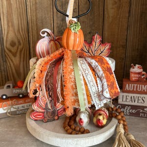 May include: A tiered tray display featuring a central orange ceramic pumpkin ornament adorned with a cascade of varied orange, brown, and cream ribbons, lace, and fabric strips, accented by two small turkey figurines and wooden bead garlands.
