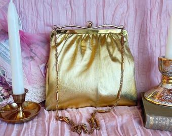 May include: A metallic gold handbag with a structured frame and a kiss-lock closure. The bag features a long, thin gold-tone chain strap with cylindrical metal accents. The material has a soft, slightly gathered texture, and the frame is finished with a polished gold-tone metal edge.