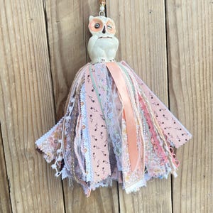 May include: A hanging ornament featuring a white ceramic owl figurine with orange accents, suspended from a floral ribbon. The owl sits atop a large, multi-layered tassel made of various fabric strips, including pink floral prints, lace, and solid peach satin ribbons.