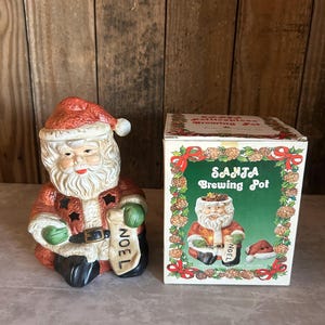 May include: A ceramic Santa Claus figurine with a red hat and coat, holding a green-gloved hand and a scroll that says "NOEL". Beside it is a box that says "SANTA Brewing Pot". The box has a Santa Claus illustration.
