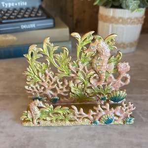 May include: A metal napkin holder featuring a detailed underwater scene with two seahorses, coral formations, and seashells. The design is finished in shades of pink, green, and gold, with small clear rhinestones embedded along the seahorses.