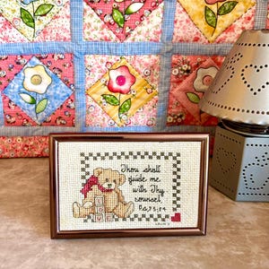 May include: A framed cross-stitch featuring a teddy bear with a red scarf and the text "Thou shalt guide me with Thy counsel, Ps.73:24." The frame is brown, and the background is a floral quilt.