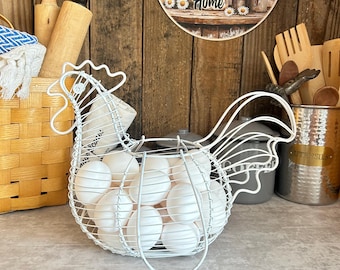 May include: A white wire egg basket shaped like a chicken, filled with numerous white eggs. The basket features a detailed wire frame forming the head, comb, and tail of the chicken, providing a sturdy structure for holding the eggs securely.