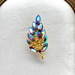 May include: A vintage gold-toned brooch in the shape of a leaf, adorned with iridescent blue and purple crystals. The brooch features a detailed design with small red accents and is set against a white background.