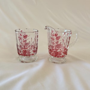 May include: Clear glass creamer and tumbler set with a red floral design. The creamer has a handle and spout, while the tumbler is a simple, straight-sided glass. Both pieces have a pedestal base and are likely vintage.