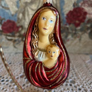 May include: A teardrop-shaped glass Christmas ornament featuring a painted depiction of a mother and child. The figure is draped in a deep red cloak with gold glitter accents, and the ornament includes a gold-colored metal cap and hanging loop.