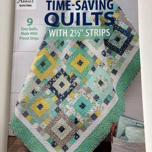 May include: A book cover titled "TIME-SAVING QUILTS" by Annie's Quilting. The cover features a colorful quilt with yellow, teal, and navy blue squares. The quilt is made with 2 1/2" strips. The book promises 9 easy quilts made with precut strips.