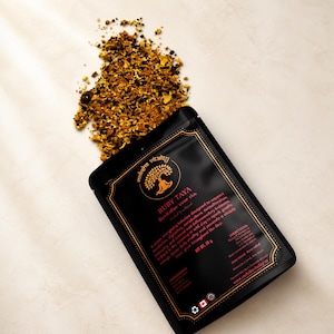 May include: A black pouch of Ruby Taya herbal tea with gold lettering and a gold border. Loose tea spills from the pouch onto a white surface. The text on the pouch reads "Revitalize Your Ihu". The net weight is 28 g.