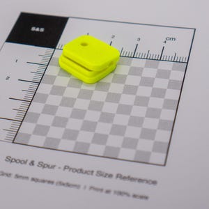May include: A bright yellow, rectangular object with a hole on top, placed on a size reference grid. The grid features a checkerboard pattern and a ruler with centimetre markings. The text "Spool & Spur - Product Size Reference" is visible.