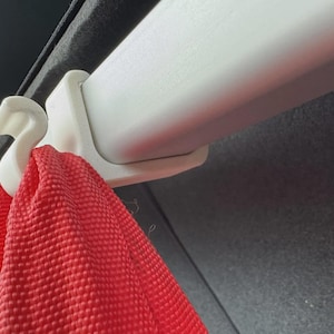 May include: Close-up of a white plastic hook and rail system, with a red woven strap hanging from the hook. The system is mounted against a dark gray surface.