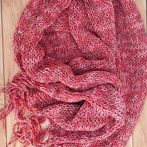Op de afbeelding: Een handgebreide sjaal in tinten rood, roze en wit, met een gefranjerde rand. De sjaal is gemaakt van een dik, getextureerd garen en is gedrapeerd op een houten oppervlak. Het ontwerp van de sjaal is een klassiek breipatroon.