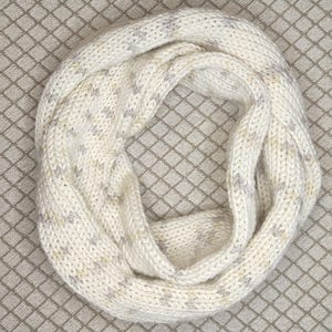 Può includere: Sciarpa a infinito color crema con trama a maglia. La sciarpa presenta un motivo maculato di macchie grigie e dorate. Il design morbido e ad anello suggerisce un accessorio accogliente e caldo, adatto al clima freddo.