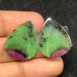 Peut inclure: Deux pierres précieuses en forme de goutte d'eau avec une surface polie. Les pierres présentent un centre vert vif, une bande magenta foncé et des inclusions noires et grises. Les pierres sont posées sur un fond sombre.
