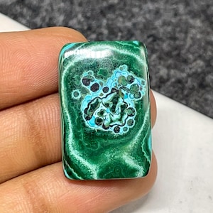 Könnte beinhalten: Ein rechteckiger, polierter Edelstein mit einem satten grünen Farbton. Der Stein weist komplizierte Muster aus hellgrünen, türkisfarbenen und schwarzen Markierungen auf. Der Edelstein wird in einer Hand gehalten, was seine natürliche Schönheit hervorhebt.