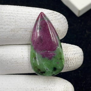 Könnte beinhalten: Ein polierter Edelstein in Tropfenform. Die obere Hälfte ist tief burgunderfarben, die an der Basis in ein leuchtendes Grün übergeht. Der Stein hat eine glatte, glänzende Oberfläche und ein natürliches, organisches Aussehen.