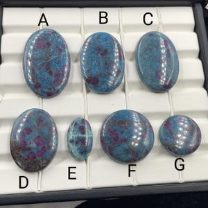 May include: A collection of polished gemstones in various shapes, including oval and round. The stones feature a teal base color with vibrant red and purple inclusions. The gemstones are displayed in a black tray with white compartments.