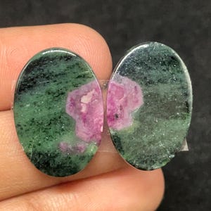 Può includere: Due pietre preziose ovali. Ogni pietra presenta una base verde con inclusioni nere e una macchia centrale di rosa vibrante. Le pietre sono lucide e hanno una superficie liscia e riflettente.