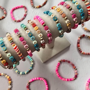 May include: A collection of colorful beaded bracelets. Some bracelets feature letter beads spelling out words, while others have striped or solid designs. The bracelets are displayed on a white stand and a light gray surface.