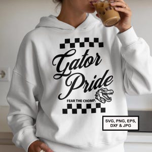 May include: White hoodie featuring the text "Gator Pride" in a black script font, with a black and white checkered pattern above and below the text. The design includes a cartoon alligator and the phrase "FEAR THE CHOMP."