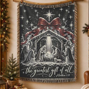 May include: A black and white woven throw blanket featuring a nativity scene under a large red bow and a bright star. The text reads, "the greatest gift of all" and "Matthew 1:21". The blanket has fringed edges and is displayed hanging from a wooden rod.