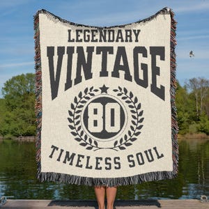 May include: A cream-colored woven blanket with black text that reads "LEGENDARY VINTAGE TIMELESS SOUL" and a circular graphic with the number "80". The blanket has a fringed edge and is held up against a lake and sky background.