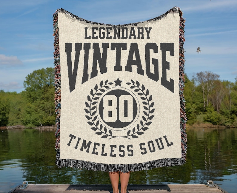 May include: A cream-colored woven blanket with black text that reads "LEGENDARY VINTAGE TIMELESS SOUL" and a circular graphic with the number "80". The blanket has a fringed edge and is held up against a lake and sky background.
