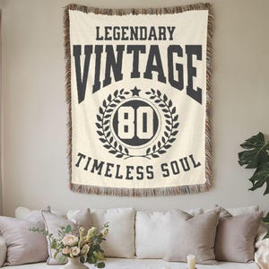 May include: Cream-colored woven throw blanket with the words "LEGENDARY VINTAGE 80 TIMELESS SOUL" in black. The blanket has a fringed edge and is hanging on a wall above a white sofa with pillows.