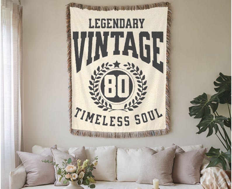 May include: Cream-colored woven throw blanket with the words "LEGENDARY VINTAGE 80 TIMELESS SOUL" in black. The blanket has a fringed edge and is hanging on a wall above a white sofa with pillows.