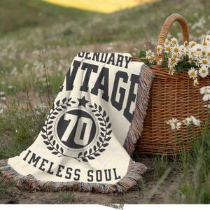 May include: A cream-colored blanket with black text reading "LEGENDARY VINTAGE 70 TIMELESS SOUL" is draped on the grass. A woven basket filled with daisies sits nearby. The blanket has a fringed edge and a star and laurel wreath design.